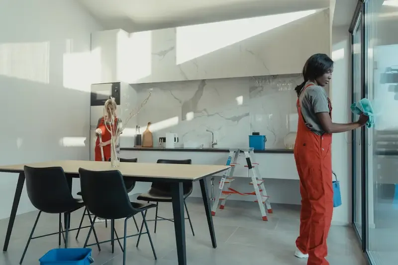 Two individuals cleaning a modern kitchen with a dining table and large windows.