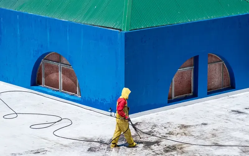 Person in yellow rain gear using a pressure washer on a blue building.